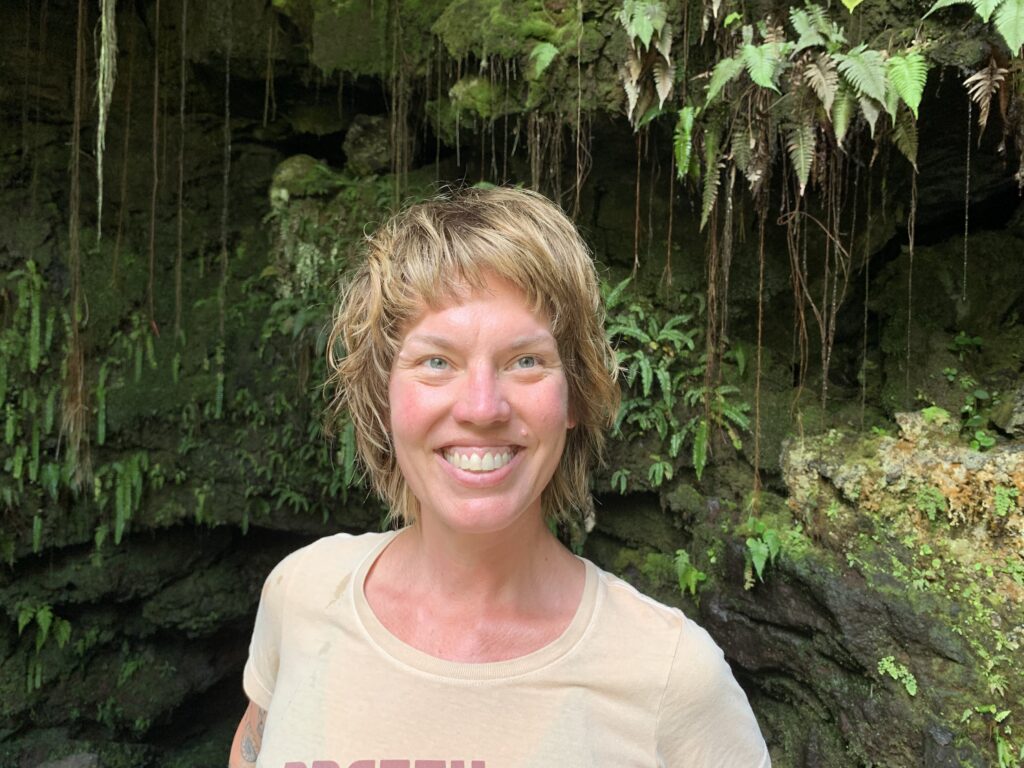 blonde woman in a light yellow tshirt against green ferns and trees