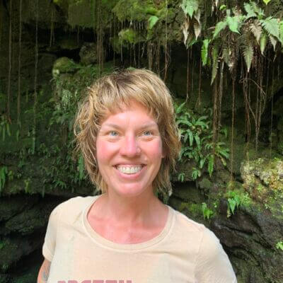 blonde woman in a light yellow tshirt against green ferns and trees