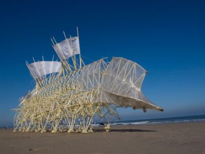 Image of large moving sculpture on beach with blue sky and tan sand.