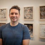 Jamie Treacy, smiling with collagraph plate and intaglio art of his students behind
